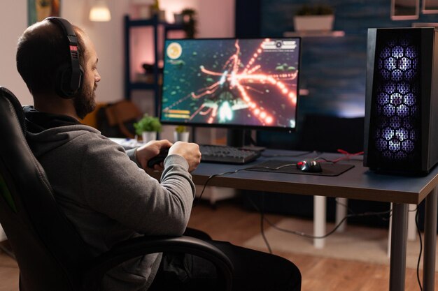 Gracz używający kontrolera do grania w gry wideo online na komputerze. Mężczyzna gra z joystickiem i słuchawkami przed monitorem. Gracz posiadający sprzęt do gier, wykonujący zabawną aktywność.