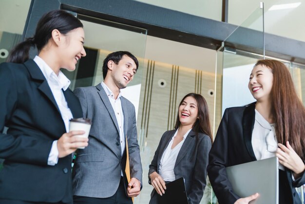 Grupa inteligentnych młodych azjatyckich biznesmenów i kobiet w formalnych strojach, przechodząc obok nowoczesnego wejścia do biura z pewnością siebie i szczęściem