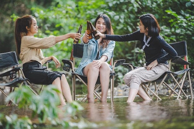 Grupa Młodych Azjatek Pije Piwo Na Swoich Krzesłach I Moczy Stopy W Strumieniu Podczas Biwakowania W Parku Przyrody. Lubią Rozmawiać I śmiać Się Razem