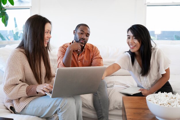 Grupa młodych ludzi korzystających razem z laptopa w domu na kanapie i jedzących przekąski