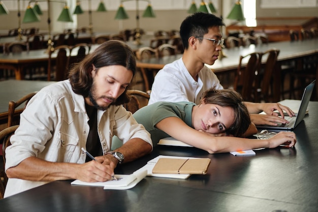 Grupa młodych międzynarodowych studentów studiujących razem w bibliotece uniwersyteckiej Atrakcyjna zmęczona studentka leżąca na stole, jednocześnie uważnie patrząc w kamerę