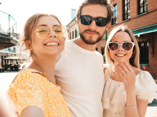 Grupa młodych trzech stylowych przyjaciół na ulicy. Mężczyzna i dwie śliczne dziewczyny ubrane w letnie ubrania. Uśmiechnięte modele bawiące się w okularach przeciwsłonecznych. Kobiety i facet robiące selfie na smartfonie