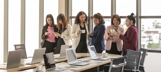 Grupa przedsiębiorców nie do poznania stojących razem w biurze w czasie łamania. praca zespołowa kobiet z relaksem i łatwością podczas korzystania ze smartfona i picia kawy.