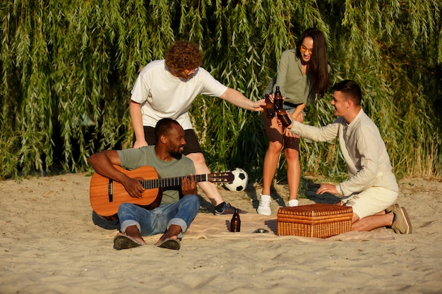 Grupa przyjaciół brzęk butelek piwa podczas pikniku na plaży. Koncepcja stylu życia, przyjaźni, zabawy, weekendu i odpoczynku.