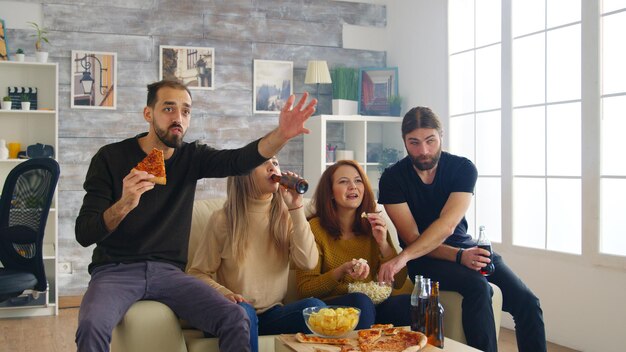 Grupa przyjaciół szczęśliwa po wygraniu mistrzostw przez ich ulubioną drużynę piłkarską. Przyjaciele siedzący na kanapie jedzący pizzę z frytkami.
