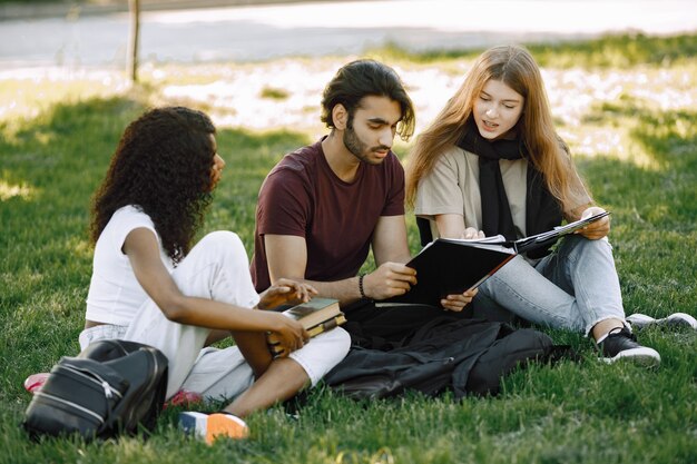 Grupa studentów międzynarodowych siedzi razem na trawie w parku na uniwersytecie. Afrykańskie i kaukaskie dziewczyny i indyjski chłopiec rozmawiający na świeżym powietrzu