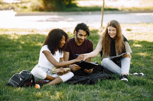 Grupa studentów międzynarodowych siedzi razem na trawie w parku na uniwersytecie. Afrykańskie i kaukaskie dziewczyny i indyjski chłopiec rozmawiający na świeżym powietrzu