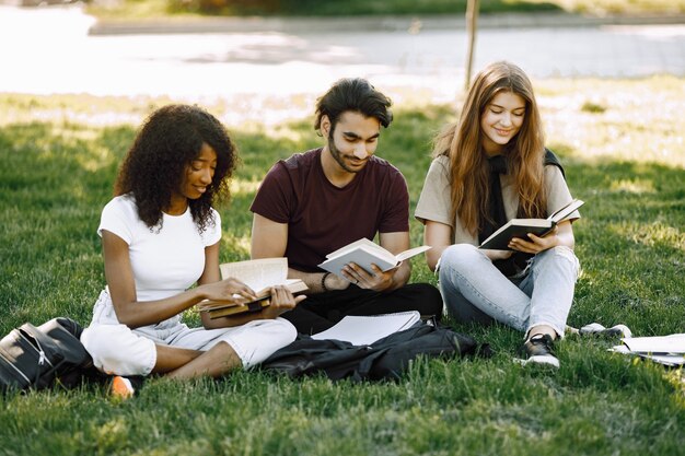 Grupa studentów międzynarodowych siedzi razem na trawie w parku na uniwersytecie. Afrykańskie i kaukaskie dziewczyny i indyjski chłopiec rozmawiający na świeżym powietrzu
