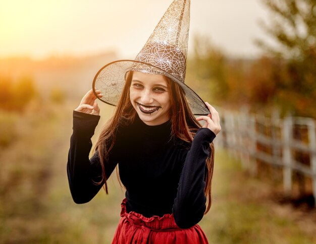 Halloweenowy portret nastoletniej dziewczyny w kapeluszu czarownicy uśmiechający się do kamery na tle przyrody