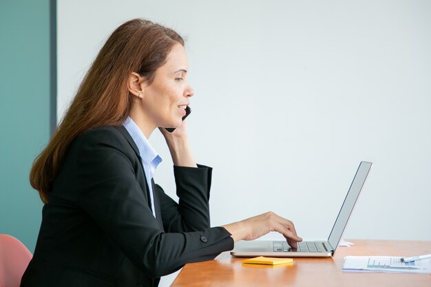 Happy całkiem młoda businesswoman rozmawia przez telefon komórkowy i uśmiecha się, pracując na komputerze w biurze, używając laptopa przy stole