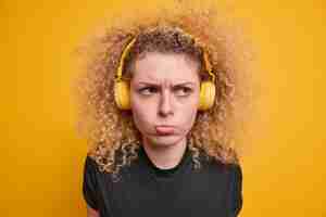 Bezpłatne zdjęcie headshot niezadowolonej kręcone włosy nastolatka ma zły nastrój dąsając się wyraz twarzy nosi bezprzewodowe słuchawki stereo słucha muzyki ubranej w dorywczo czarną koszulkę na białym tle nad żółtą ścianą