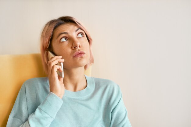 Headshot pięknej nastolatki z kolczykiem w nosie patrząc w górę z zamyślonym wyrazem twarzy podczas rozmowy przez telefon komórkowy, próbując coś sobie przypomnieć.