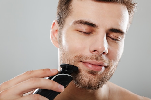 Headshot rasy kaukaskiej zadowolony facet 30s rozbierany w łazience podczas golenia twarzy trymerem na szarej ścianie