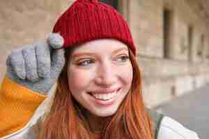 Bezpłatne zdjęcie headshot szczęśliwej rudej dziewczyny z piegami nosi czerwony kapelusz i rękawiczki zimą spaceruje po mieście