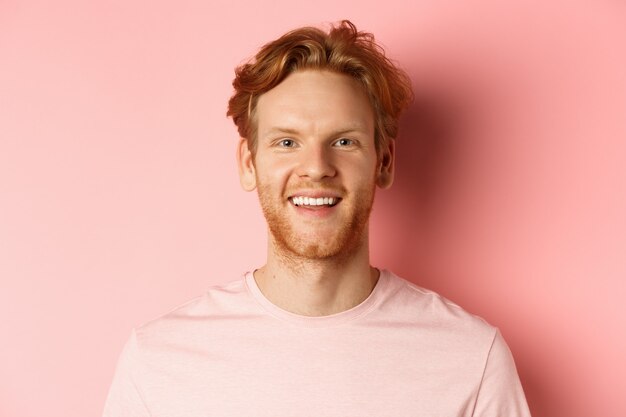 Headshot szczęśliwy rudy mężczyzna z brodą i białymi zębami, uśmiechnięty podekscytowany do kamery, stojący na różowym tle.