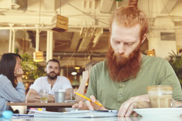 Hipster freelancer trzymający ołówek, robiąc notatki na kartkach z grafiką, wykorzystujący cyfrowy tablet do zdalnej pracy w przestrzeni coworkingowej.