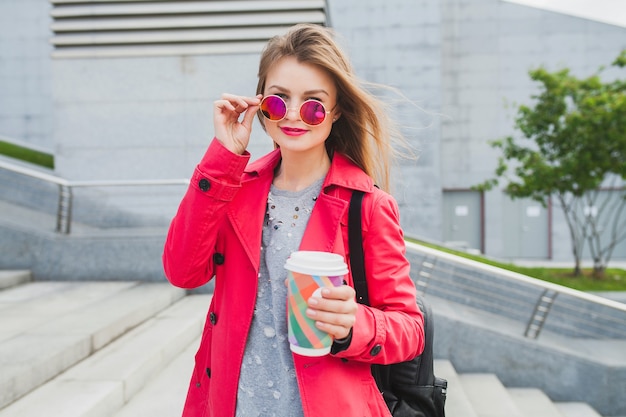Hipster młoda kobieta w różowy płaszcz, dżinsy na ulicy z plecakiem i kawą, słuchanie muzyki na słuchawkach