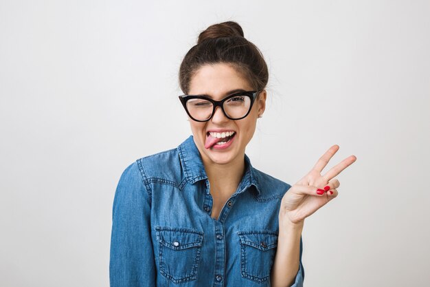 Hipster młoda stylowa kobieta w modnych okularach i dżinsowej koszuli robi zabawny wyraz twarzy, pokazując język, mrugając, gest znaku pokoju, pozytywny