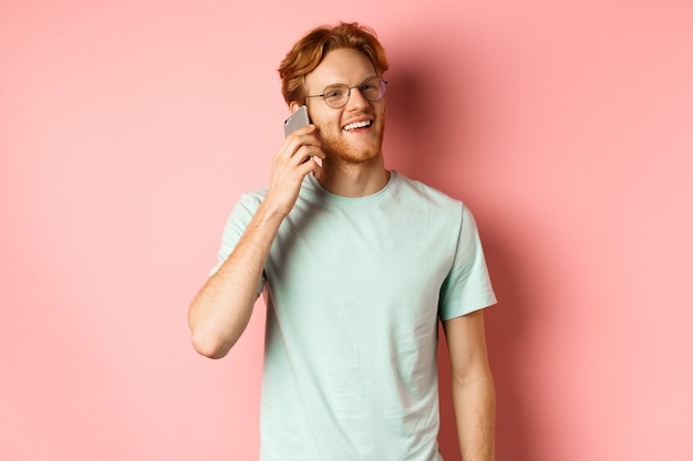 Hipster przystojny facet z rudymi włosami i brodą rozmawia przez telefon komórkowy, dzwoni do kogoś i wygląda szczęśliwy, stojąc na różowym tle.