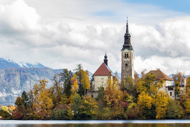 Bezpłatny plik Zdjęcie historyczny kasztel otaczający zielonymi drzewami blisko jeziora pod białymi chmurami w krwawiący, slovenia