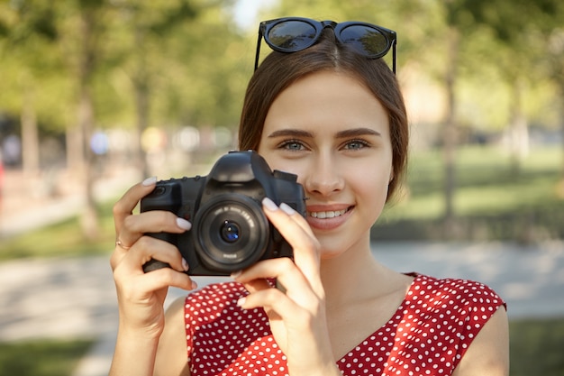 Hobby, rozrywka, zawód i koncepcja czasu letniego. Urocza szczęśliwa młoda studentka robi zdjęcia ludzi i przyrody w parku przy użyciu aparatu DSLR, uśmiechnięta, o radosnym wyrazie twarzy