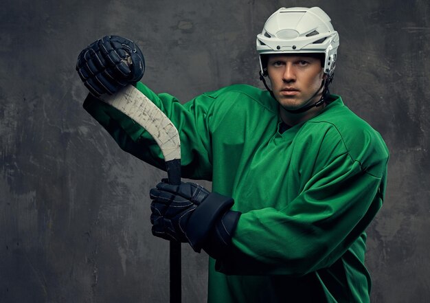 Hokeista ubrany w zielony ochronny sprzęt i biały kask stojący z kijem hokejowym na szarym tle.