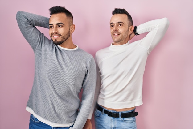 Homoseksualna para stojąca na różowym tle uśmiechająca się pewnie dotykająca włosów gestem ręki w górę, pozująca atrakcyjnie i modnie