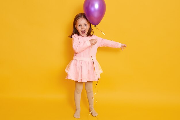 Horyzontalny strzał mała dziewczynka z purpurowym balonem odizolowywającym nad żółtym wszystkie żeńskie dziecko krzyczy coś, świętuje herbirtday, dzieciak jest ubranym różową suknię i ma ciemne włosy.