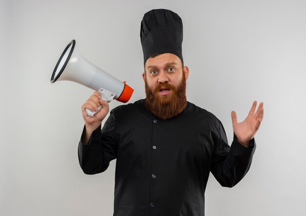 Imponujący młody przystojny kucharz w mundurze szefa kuchni trzymający głośnik i podnoszący rękę na białym tle na białej ścianie