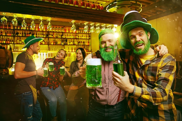 Impreza z okazji Dnia Świętego Patryka. Szczęśliwi przyjaciele świętują i piją zielone piwo. Młodzi mężczyźni i kobiety w zielonych kapeluszach. Wnętrze pubu.