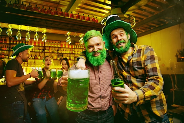 Impreza z okazji Dnia Świętego Patryka. Szczęśliwi przyjaciele świętują i piją zielone piwo. Młodzi mężczyźni i kobiety w zielonych kapeluszach. Wnętrze pubu.