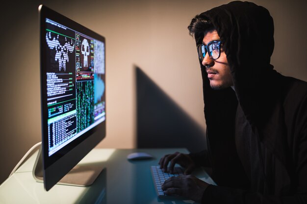 Indianmale haker ze smartfonem i kodowaniem na ekranie komputera w ciemnym pokoju