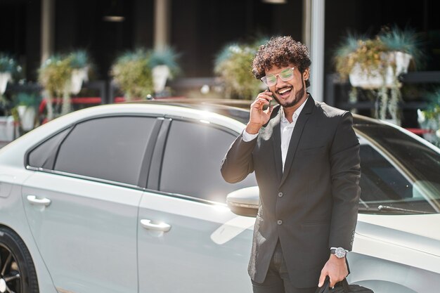 Indyjski biznesmen z kręconymi włosami rozmawia przez telefon przed samochodem