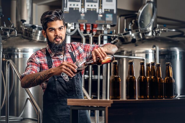 Indyjski brodaty wytatuowany hipster męski producent degustujący i prezentujący piwo rzemieślnicze w minibrowarze.