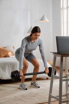 Instruktorka fitness podnosząca ciężary w domu przed laptopem