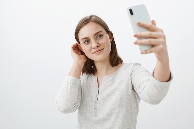 Inteligentna i czuła kobieta w okularach, strzepująca pasmo włosów za uchem i uśmiechająca się uroczo podczas robienia selfie na nowym smartfonie, pozująca na szarej ścianie, zadowolona z robienia postów w sieci społecznościowej