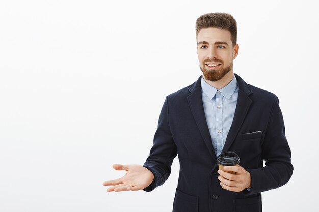 Inteligentny i kreatywny charyzmatyczny przedsiębiorca męski w stylowym garniturze trzymający papierowy kubek z kawą podczas przerwy rozmawiający z partnerem biznesowym, omawiający pracę i pieniądze, gestykulujący z uśmiechem na dłoni pewny