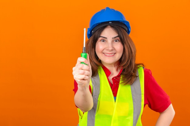 Inżynier kobieta ubrana w kamizelkę budowlaną i hełm ochronny stojący ze śrubokrętem uśmiechnięty przyjazny na odosobnionej pomarańczowej ścianie