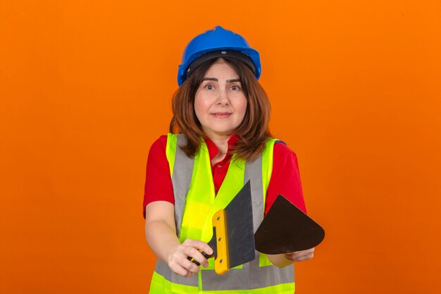 Inżynier kobieta ubrana w kamizelkę budowlaną i hełm ochronny wyciągająca kielnię i szpachlę w dłoniach wyglądająca na zdezorientowaną na odizolowanej pomarańczowej ścianie