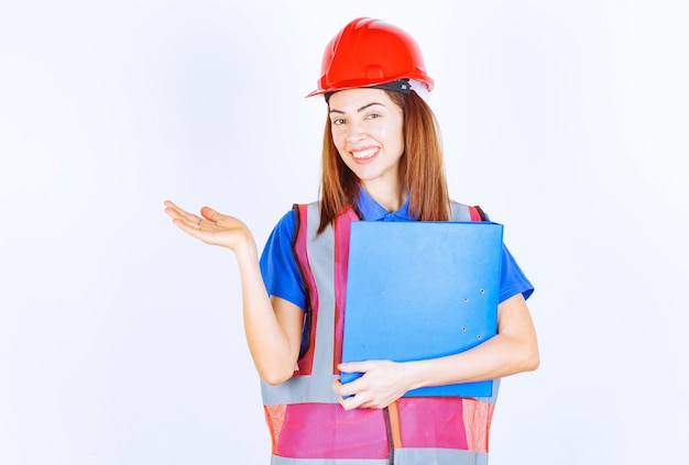 Inżynierka w czerwonym hełmie trzymająca niebieski folder projektu i przedstawiająca kogoś lub coś.