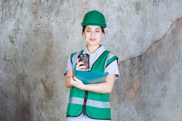 Inżynierka w zielonym mundurze i kasku, trzymająca czarną filiżankę kawy i niebieski folder projektu.
