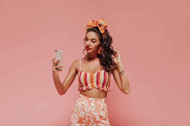 Jasno opalona kobieta z pomarańczową chustką i stylowymi kolczykami w pasiastym topie i różowych spodniach z nadrukiem robi selfie na izolowanej ścianie