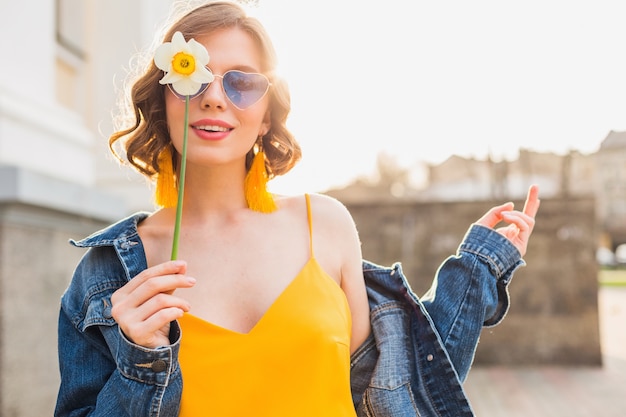 Jasny portret pięknej kobiety trzymającej kwiat, żółta sukienka, kurtka dżinsowa, styl hipster, letni trend w modzie, uśmiech, modne okulary przeciwsłoneczne