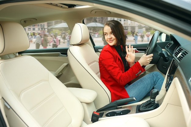 Jazda po mieście. Młoda atrakcyjna kobieta podczas prowadzenia samochodu. Młody całkiem kaukaski model w eleganckiej stylowej czerwonej kurtce siedzi w nowoczesnym wnętrzu pojazdu