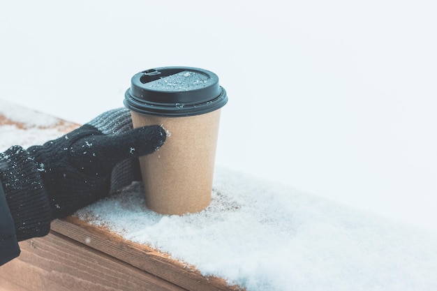 Jedna jednorazowa filiżanka kawy na śniegu na drewnianej desce. ręka w dzianej czarnej rękawiczce trzyma paczkę