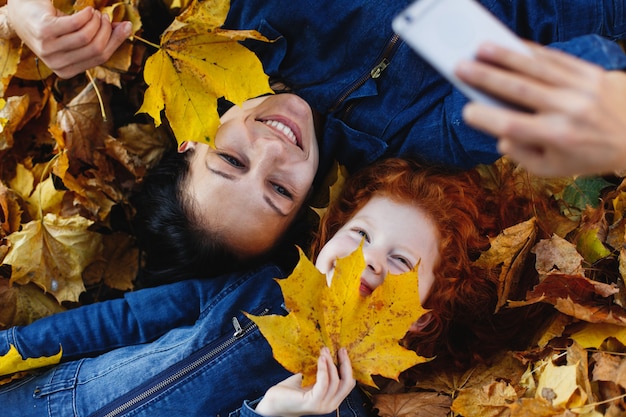 Jesienne wibracje, portret rodzinny. Urocza mama i jej córeczka z rudymi włosami bawią się, biorąc selfie na sm