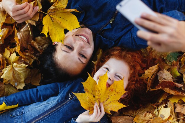 Jesienne wibracje, portret rodzinny. Urocza mama i jej córeczka z rudymi włosami bawią się, biorąc selfie na sm