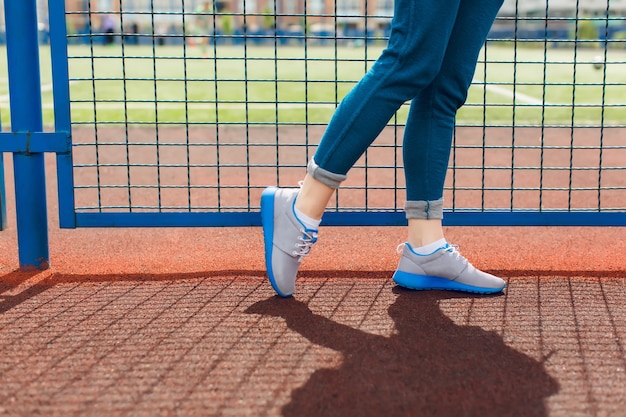 Bezpłatne zdjęcie jest to zdjęcie nóg dziewczyny idącej w pobliżu niebieskiego płotu na stadionie. nosi szare trampki z niebieską linią i niebieskie spodnie.