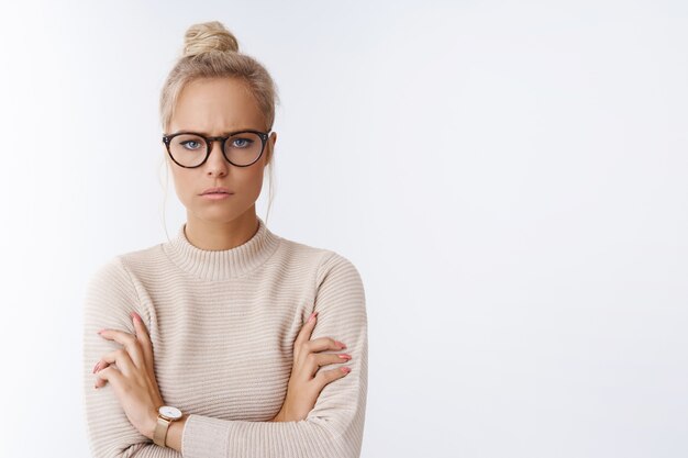 Jestem na ciebie wściekły. Portret dąsającej się niezadowolonej i dziecinnej ślicznej blond kobiety z kok fryzurowymi okularami krzyżującymi ręce nad klatką piersiową nastrojowy i agresywny marszczący brwi, dąsający się zły na białej ścianie