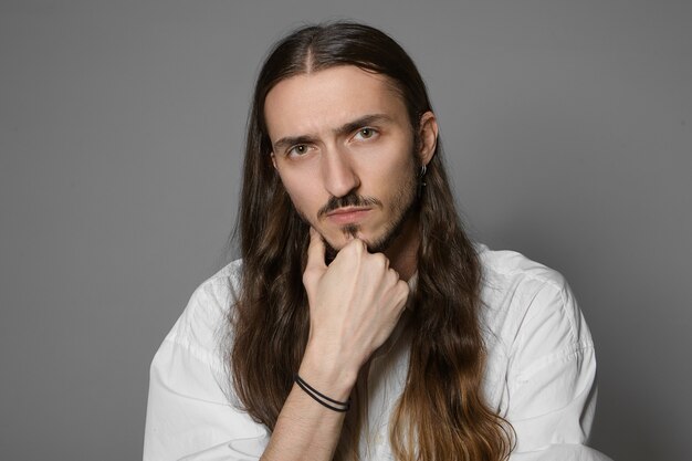 Język ciała. Portret przystojnego, nieogolonego młodego Europejczyka z wąsami i długą luźną fryzurą dotykającego brody, zastanawiającego się nad jakimś pomysłem, problemem lub projektem, pozowanie na białym tle